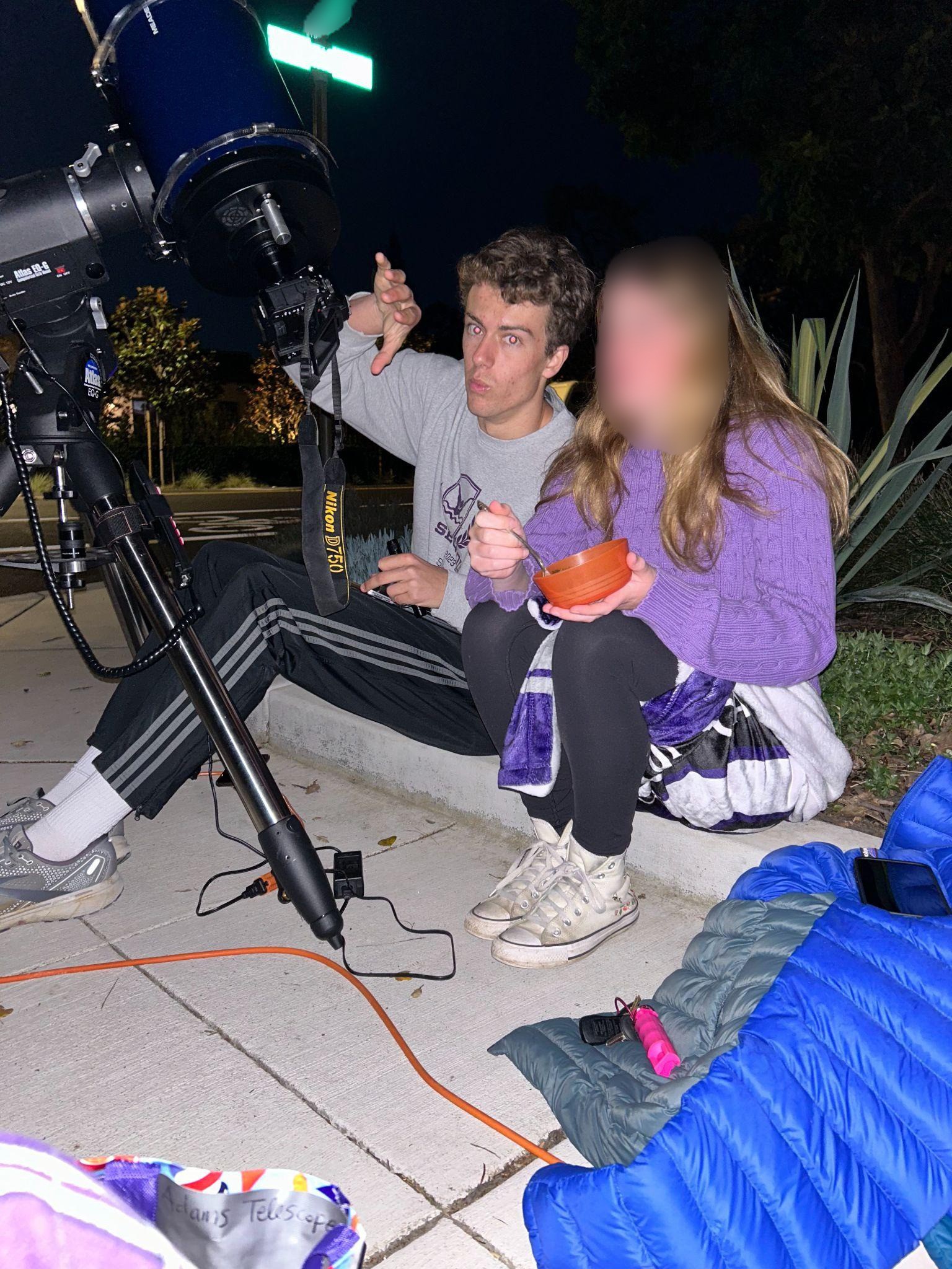 Two people sitting beside a telescope and DSLR at night