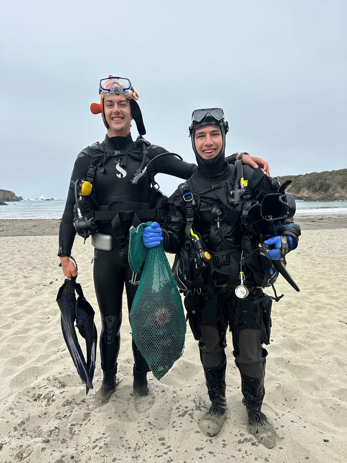 Two divers on the beach after a dive, holding fins and a mesh bag of urchins